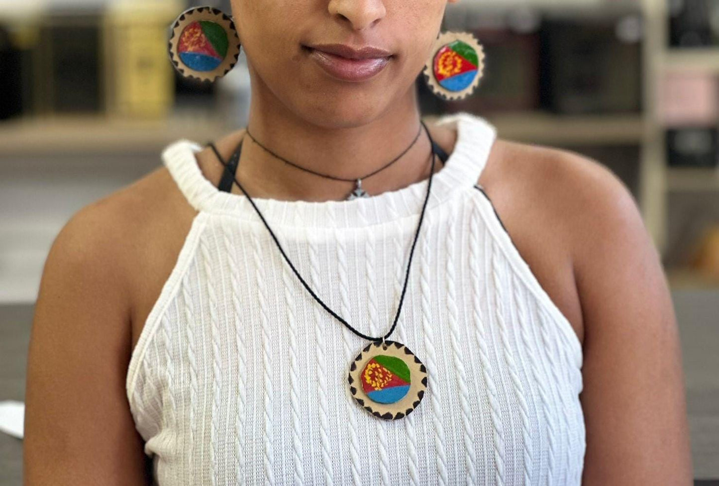 A person wearing a white knit top and a necklace with an Eritrean flag-inspired pendant. They are also wearing large, circular earrings with the same Eritrean flag colors.