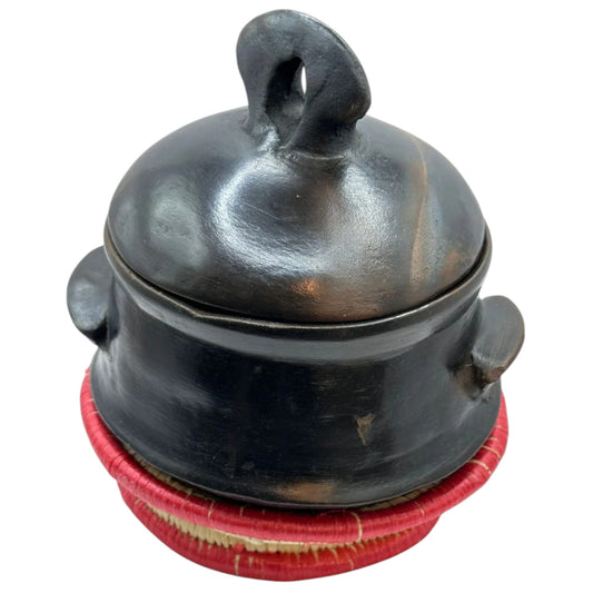 A traditional Habesha clay pottery stew bowl, known as Shekla, with a black and brown color and a hand holding palm leaf on the lid.